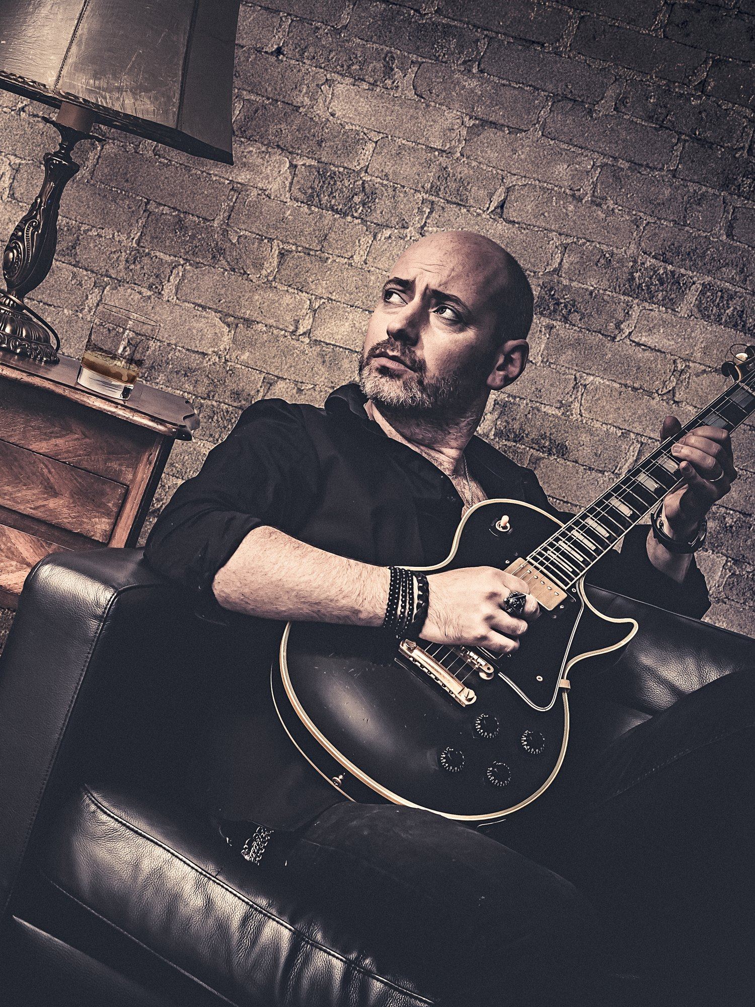 Quentin Piepszownik assis dans un canapé en cuir, une guitare électrique noire en main, le regard levé.