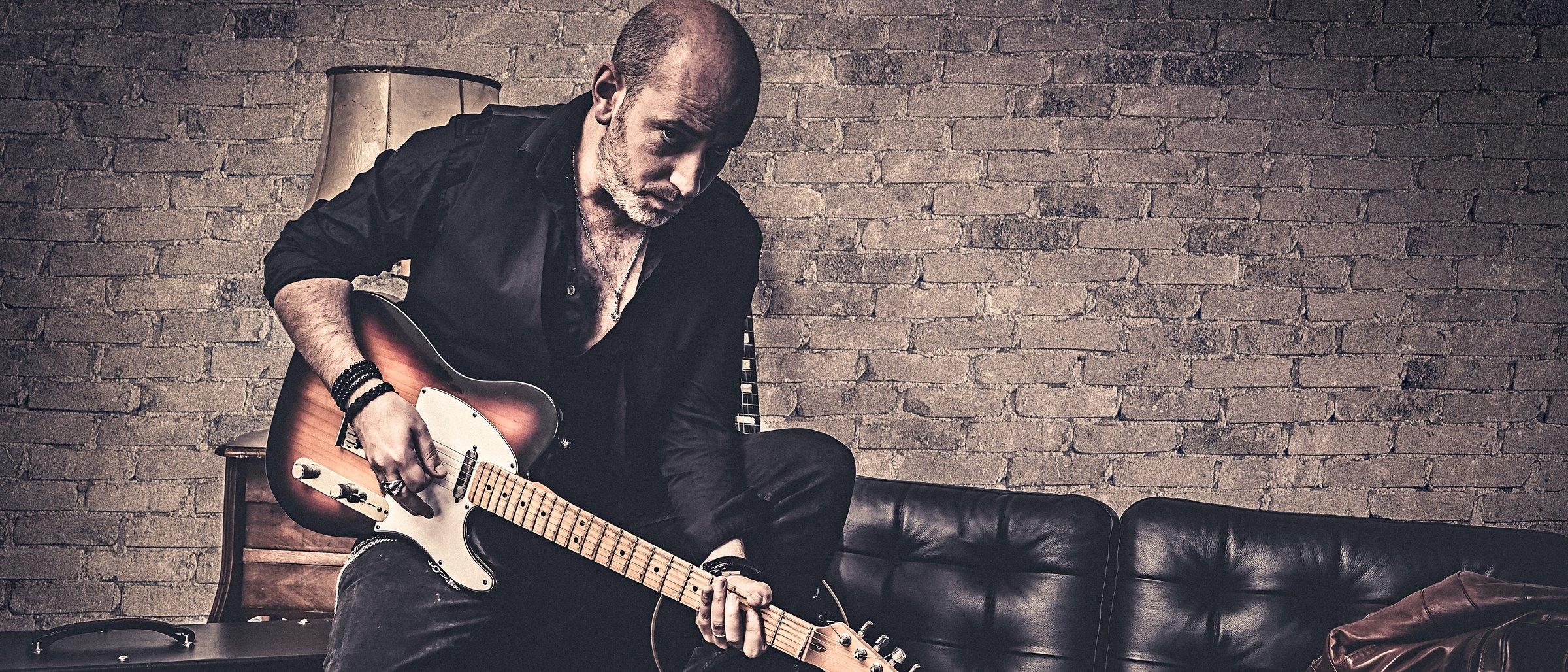 Quentin Piepszownik penché sur une guitare électrique sunburst, devant un mur de briques.
