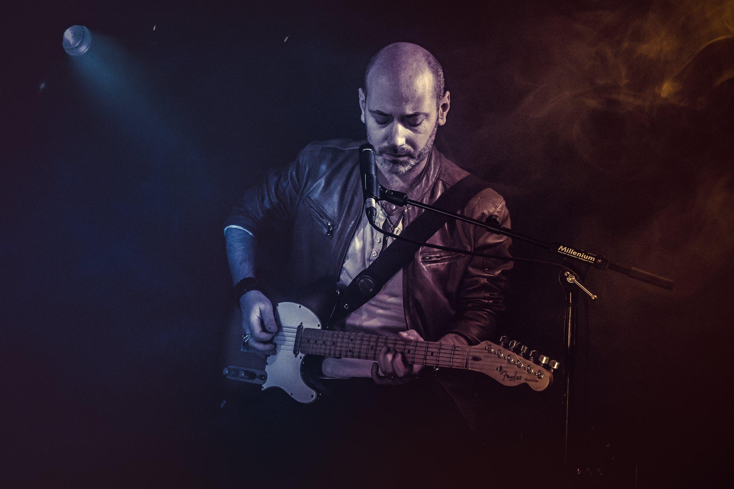 Quentin Piepszownik joue de la guitare électrique sur scène, dans un faisceau de lumière bleue.